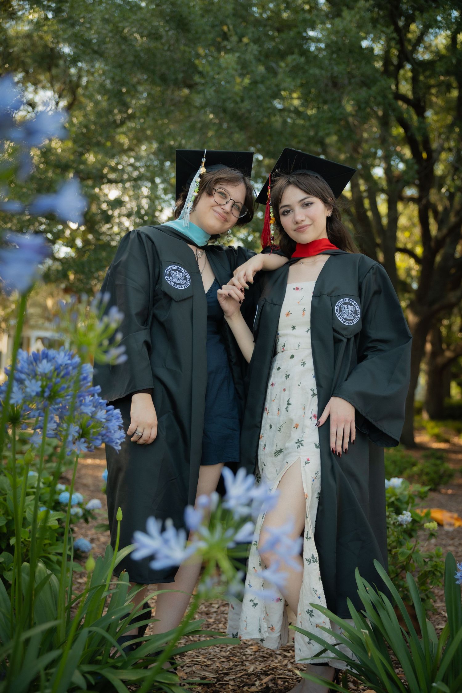 Two female graduates in dark green caps and gowns pose among blue flowers and lush garden greenery.