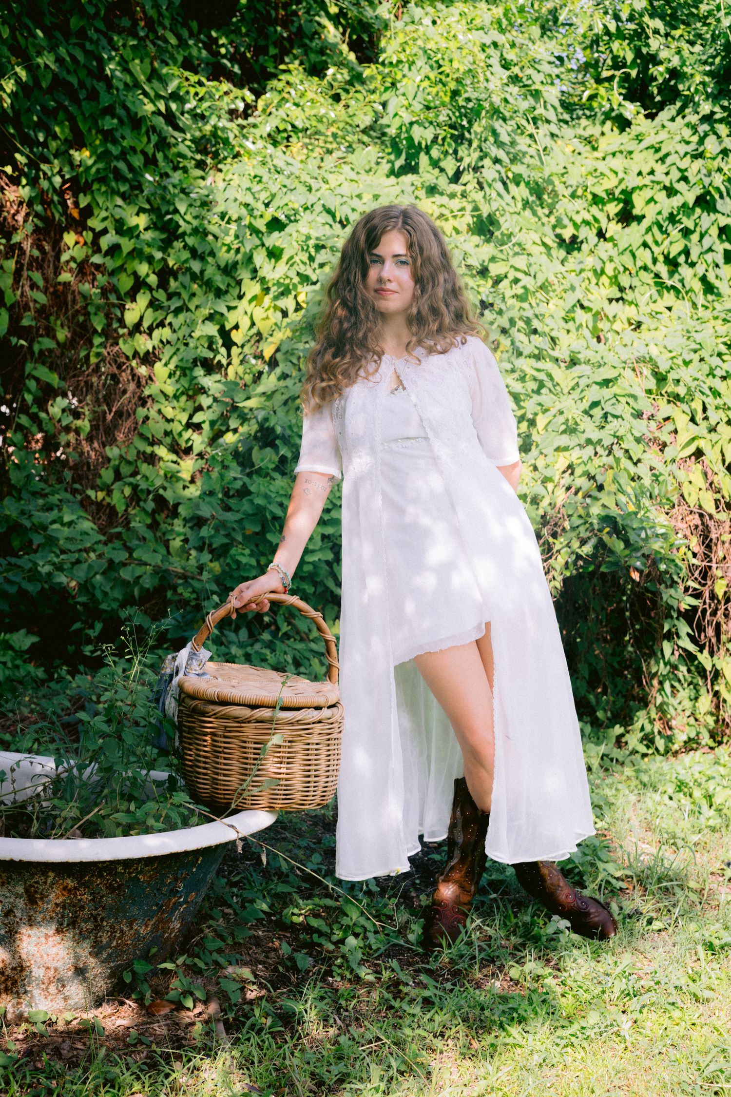 Woman in white dress holding wicker basket standing in lush green garden wearing brown boots.
