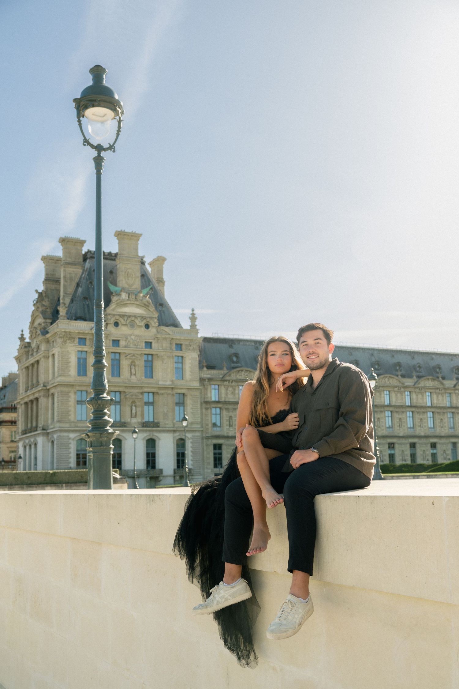 Couple sitting closely together on a stone ledge with the Louvre Museum in the background.