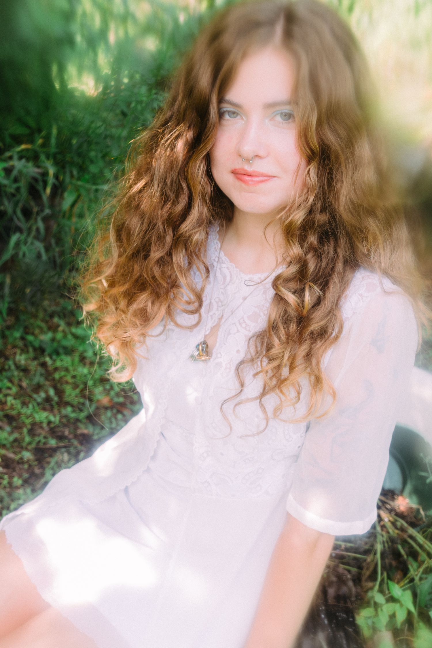 Young woman with long curly auburn hair wearing a white lace dress, sitting outdoors among green foliage.