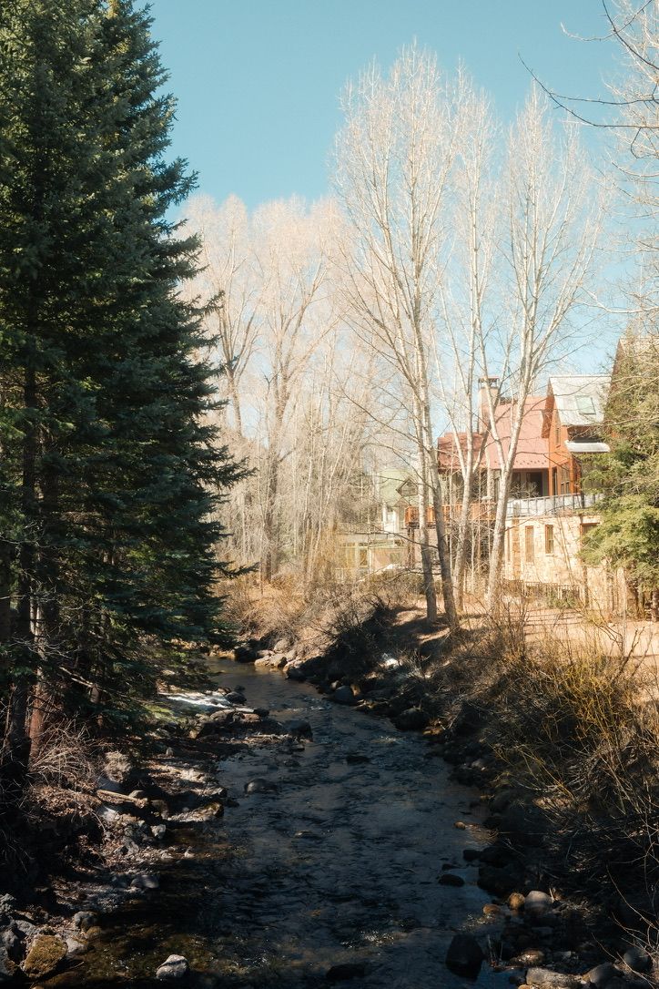 Rocky mountain stream winding through bare trees with rustic red cabin in background.