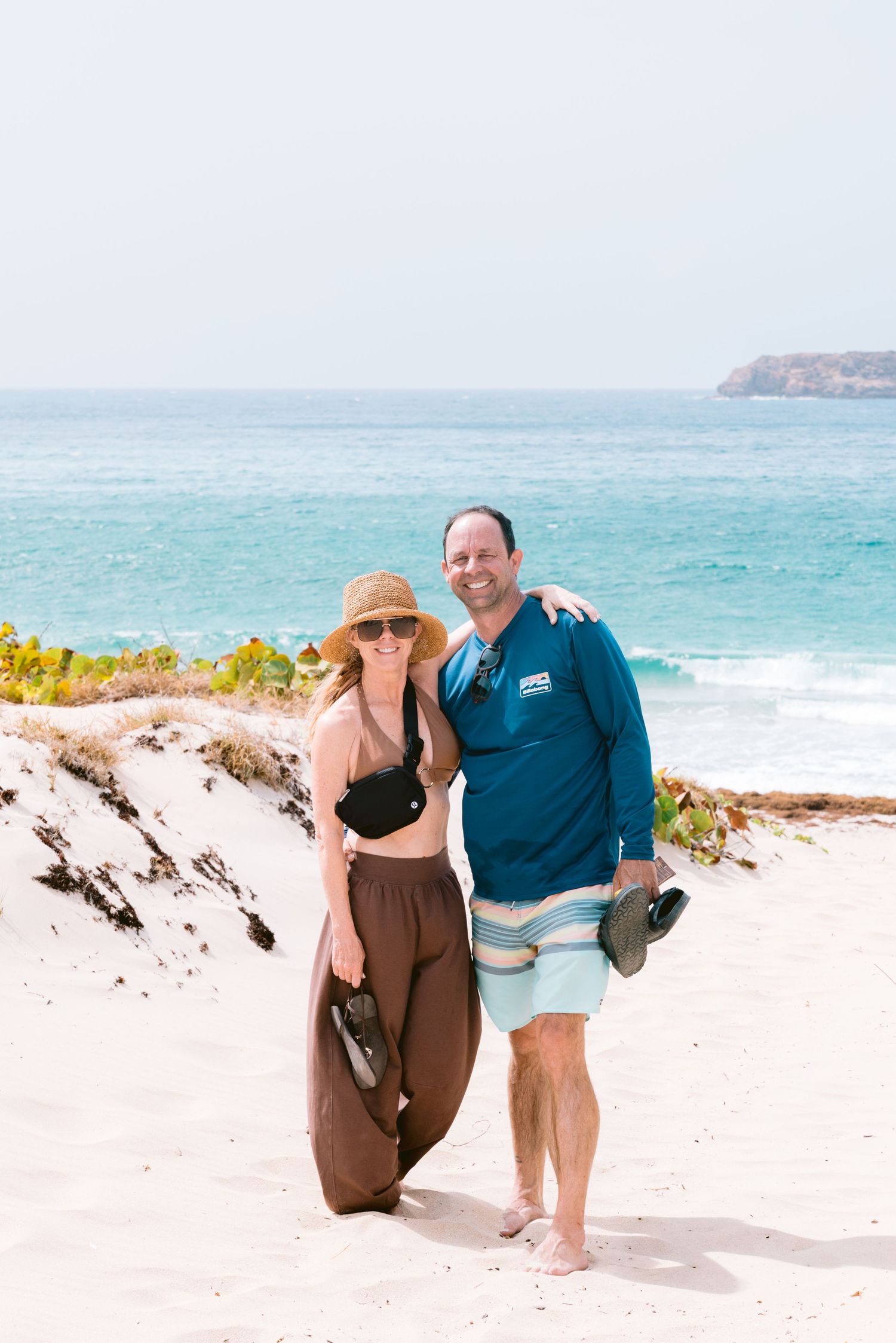 Couple smiling on white sand beach with turquoise ocean waves and rocky headland in background on sunny day.