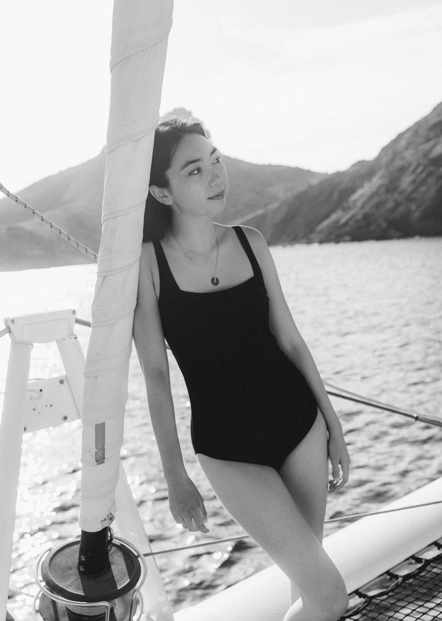 Black and white photo of a young woman in a swimsuit posing on a sailboat with mountains and water in the background.