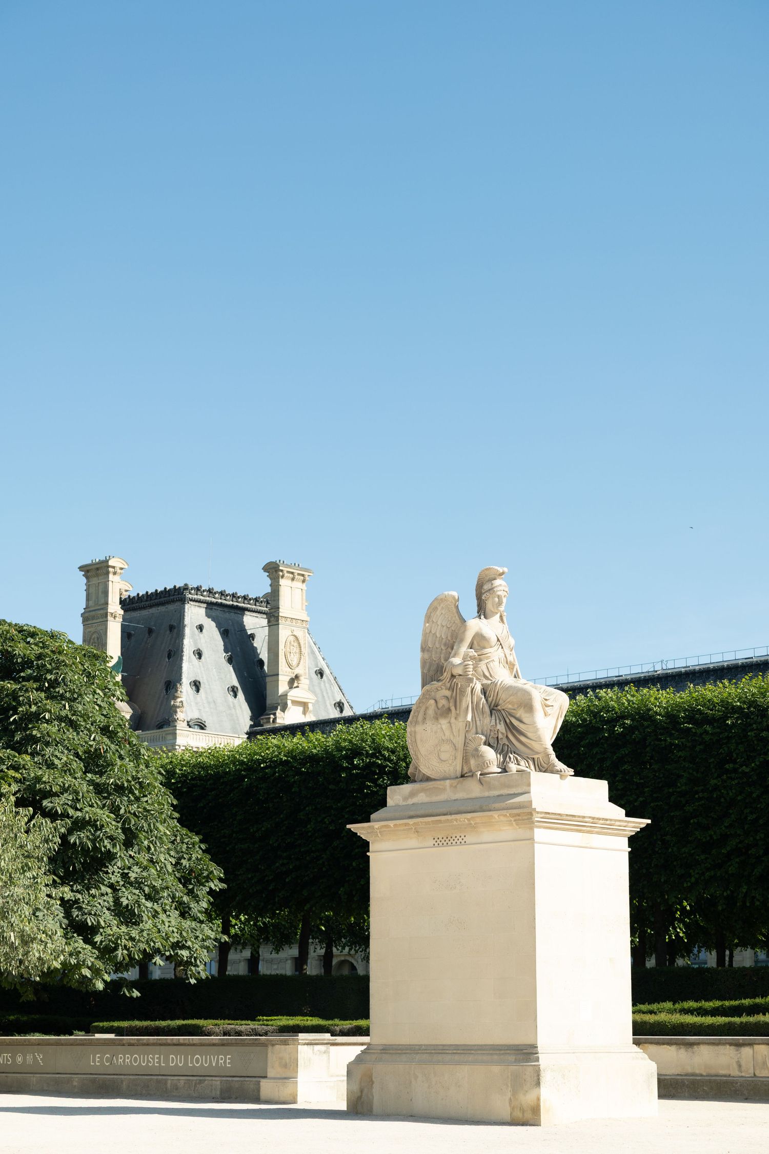 A winged sphinx statue stands in the Tuileries Garden with the Louvre museum visible behind green hedges.