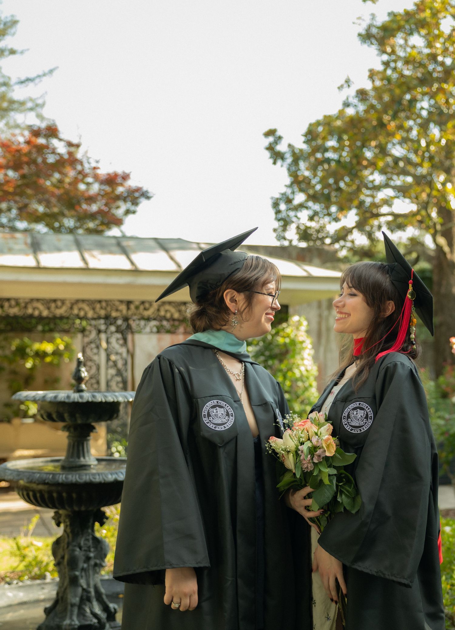Two SCAD graduates in black robes smile at each other near stone fountain holding pink roses.