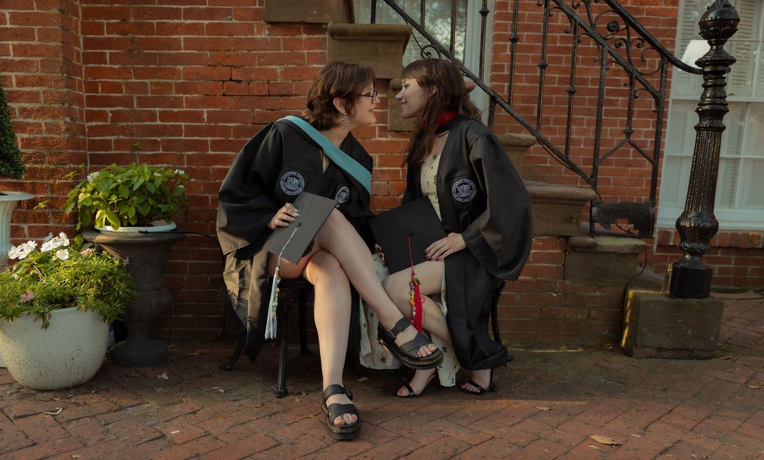 Two graduates in black robes share a kiss on brick steps outside a red brick building.