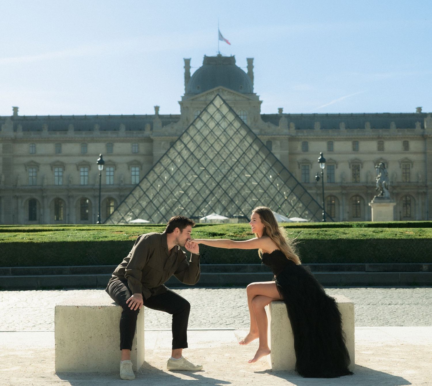 Couple sitting on stone benches facing each other with the Louvre Pyramid in the background, Paris.