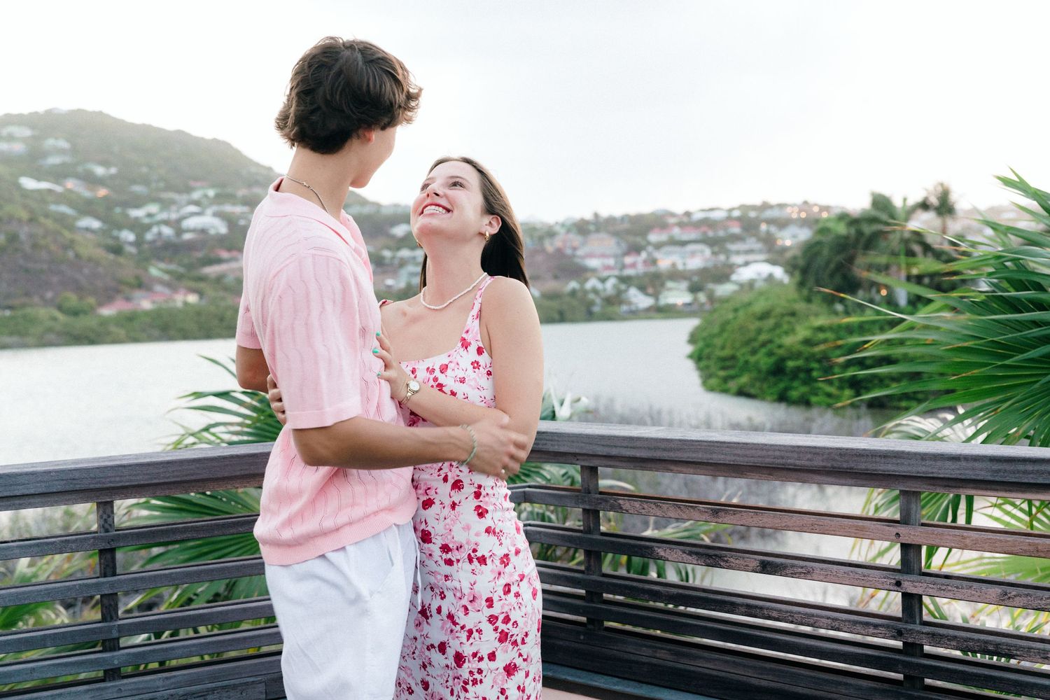 Couple embracing on a balcony overlooking a tropical waterfront, smiling at each other in casual summer attire.