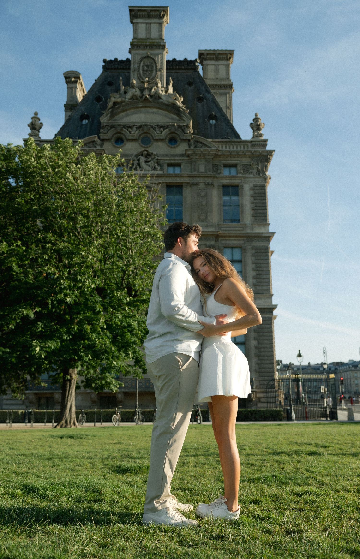 Couple embracing tenderly in white outfits on lawn before historic Paris building.
