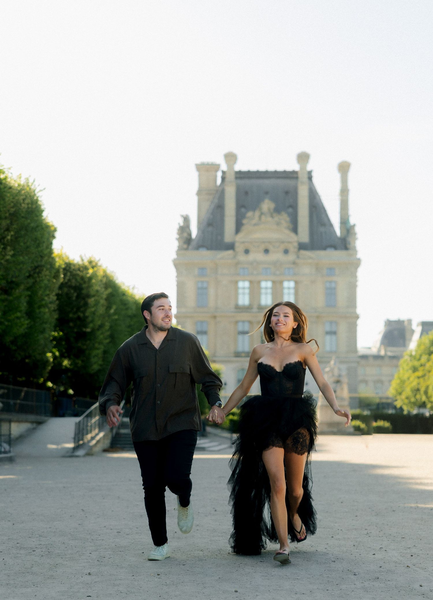 Couple in black and olive outfits running hand in hand near Louvre Paris.