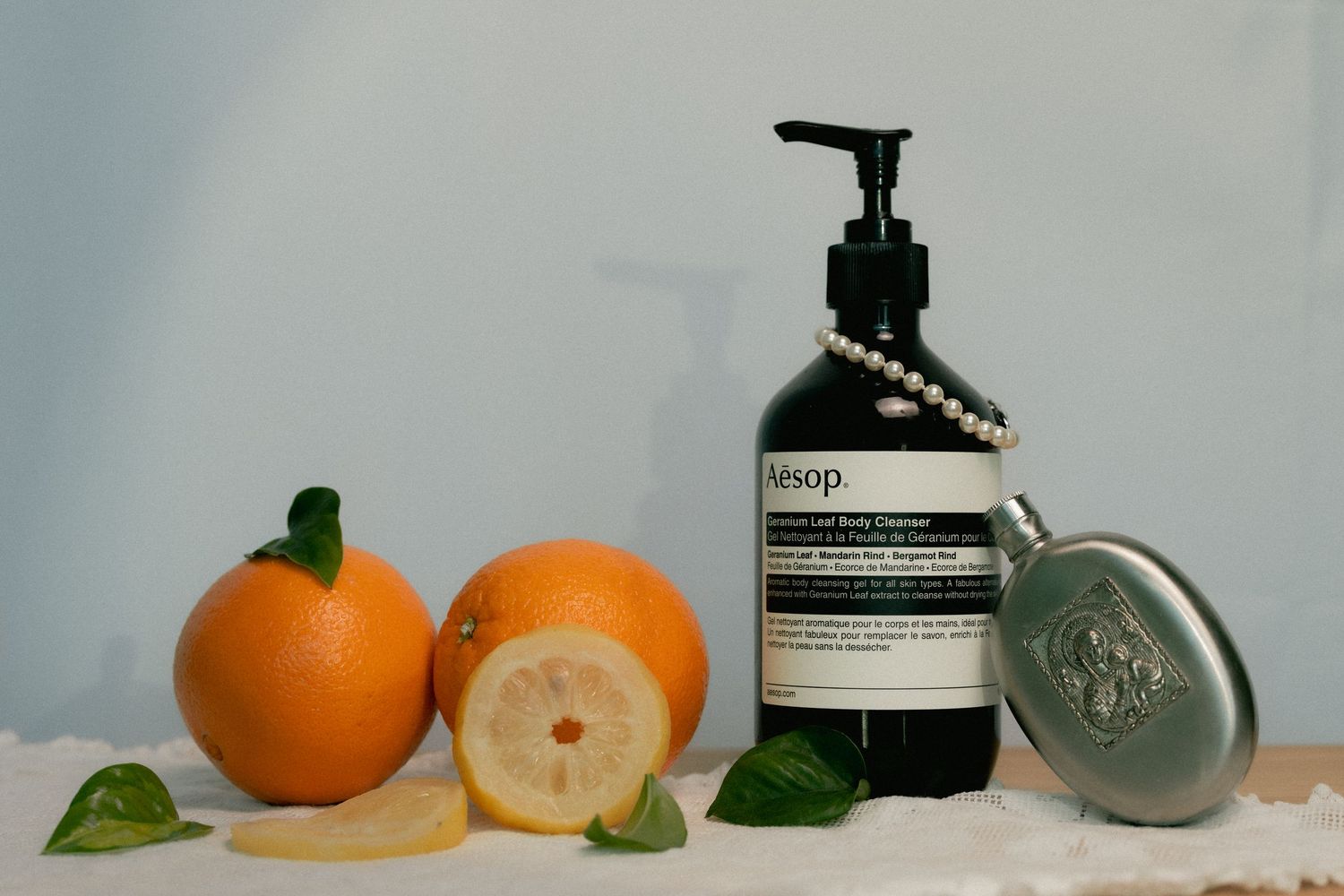 Aesop body cleanser pump bottle displayed with fresh oranges, lemon slices, green leaves, and a silver flask on a light blue background.