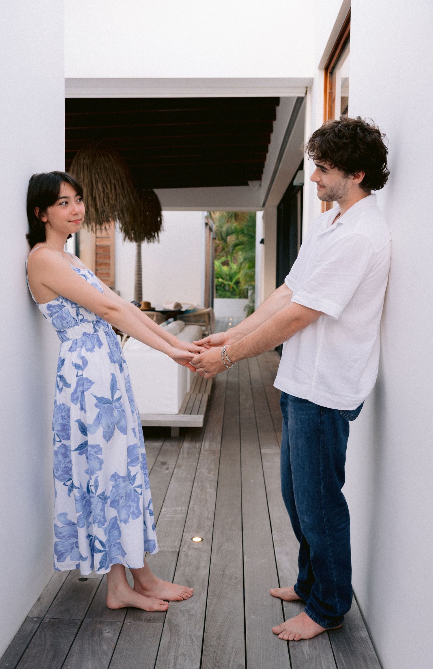 A couple holds hands barefoot on a wooden deck, woman in blue floral dress, man in white shirt and jeans.