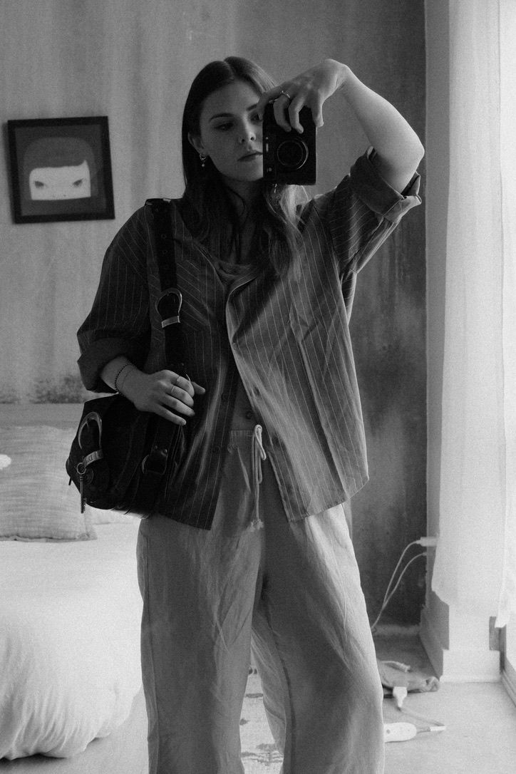 Black and white mirror selfie of a woman in linen outfit holding a camera with a bag over her shoulder.