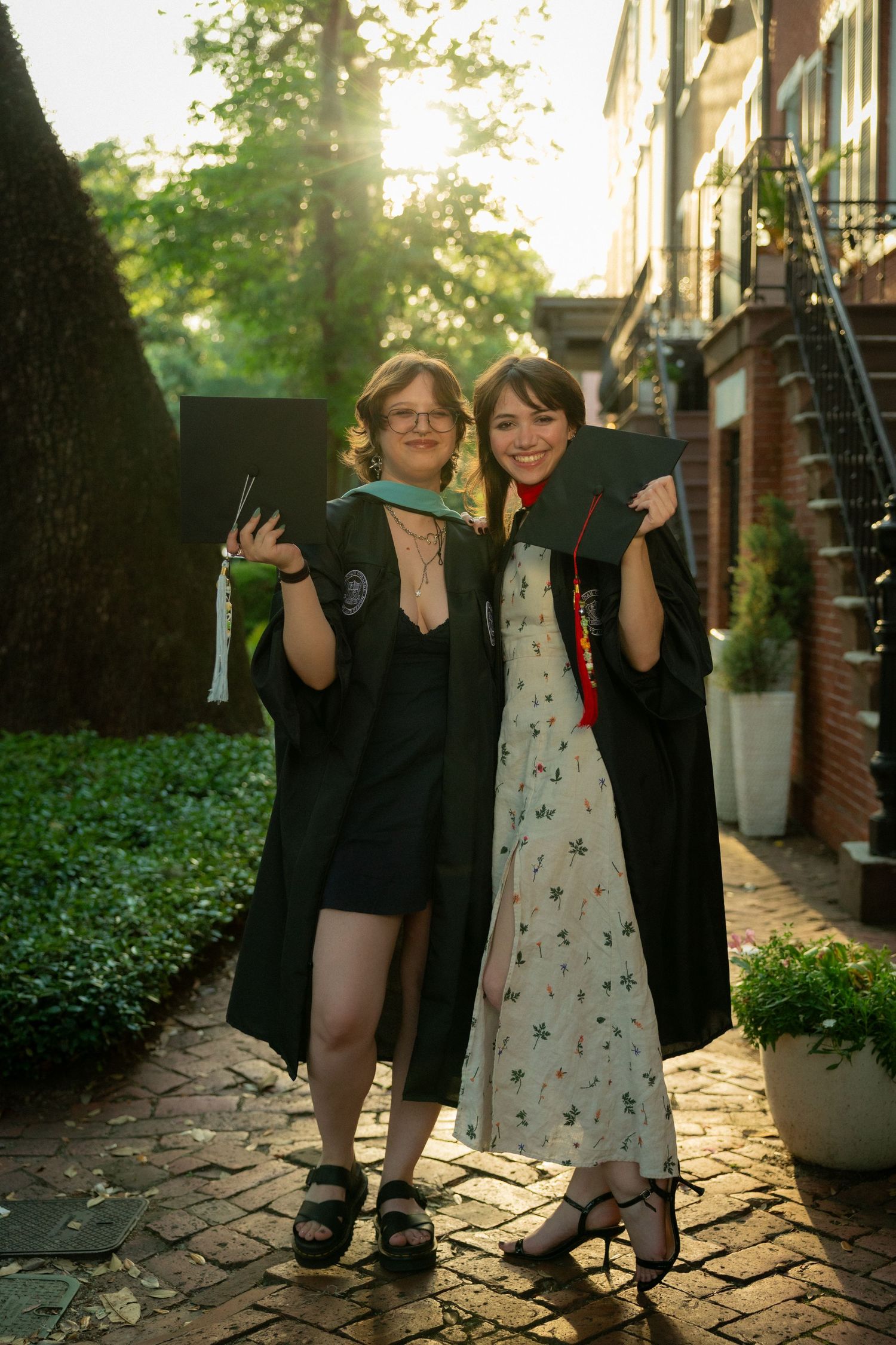 Two graduates in black gowns pose with caps on sunny brick path.
