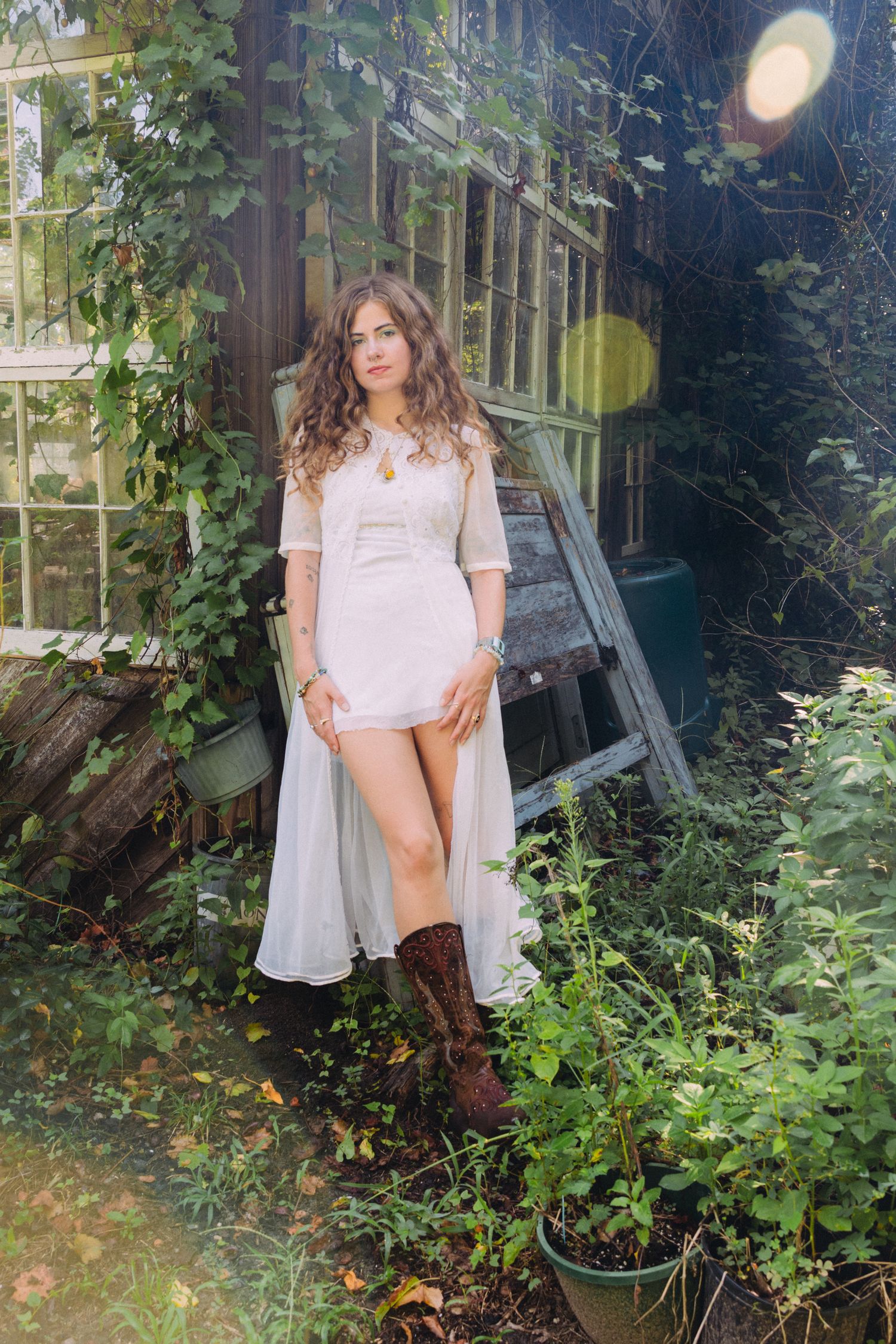 Woman in white dress and brown boots posing in a lush garden near a rustic wooden shed.