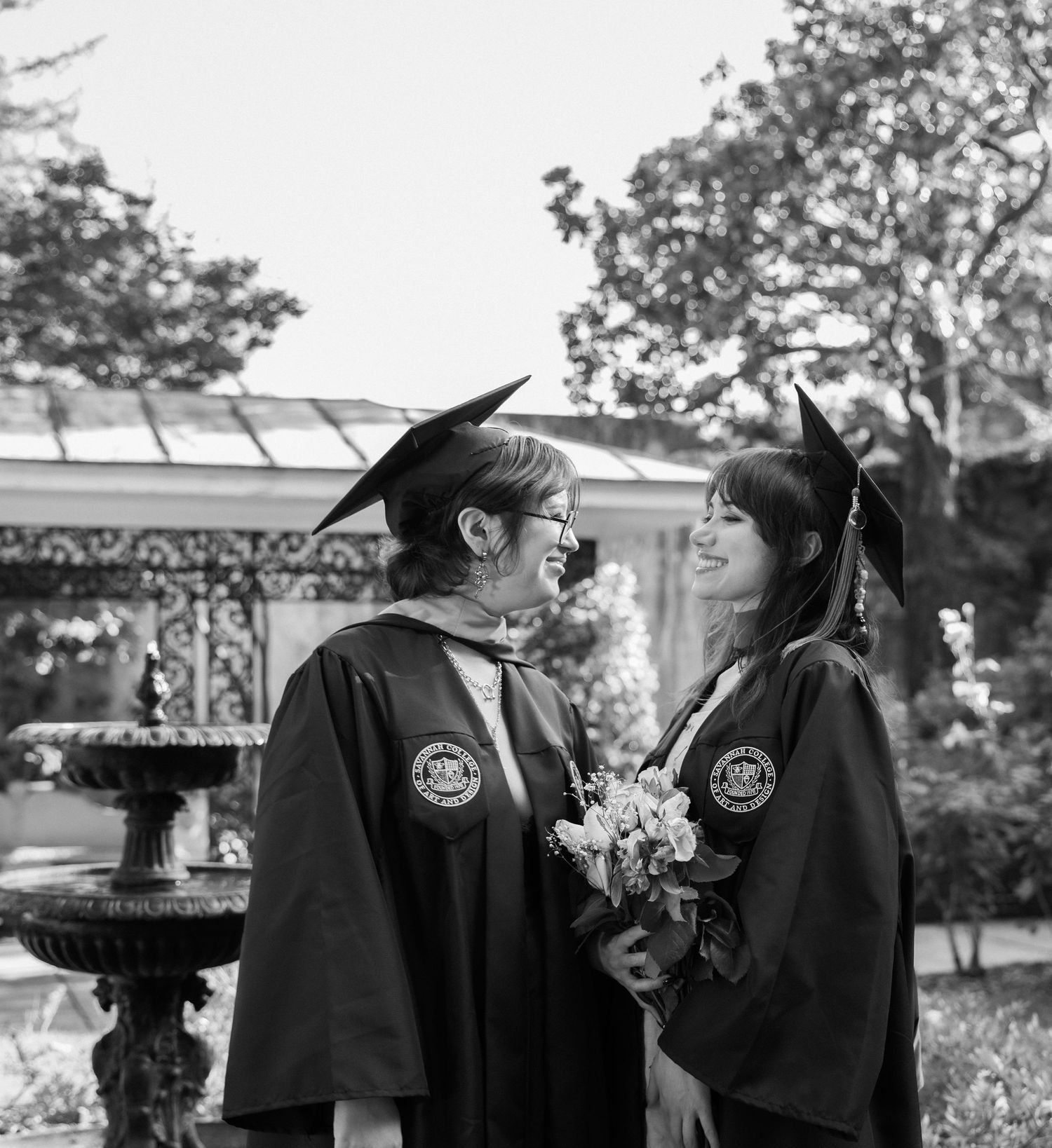 Two smiling graduates in black caps and gowns face each other near a fountain in black and white.