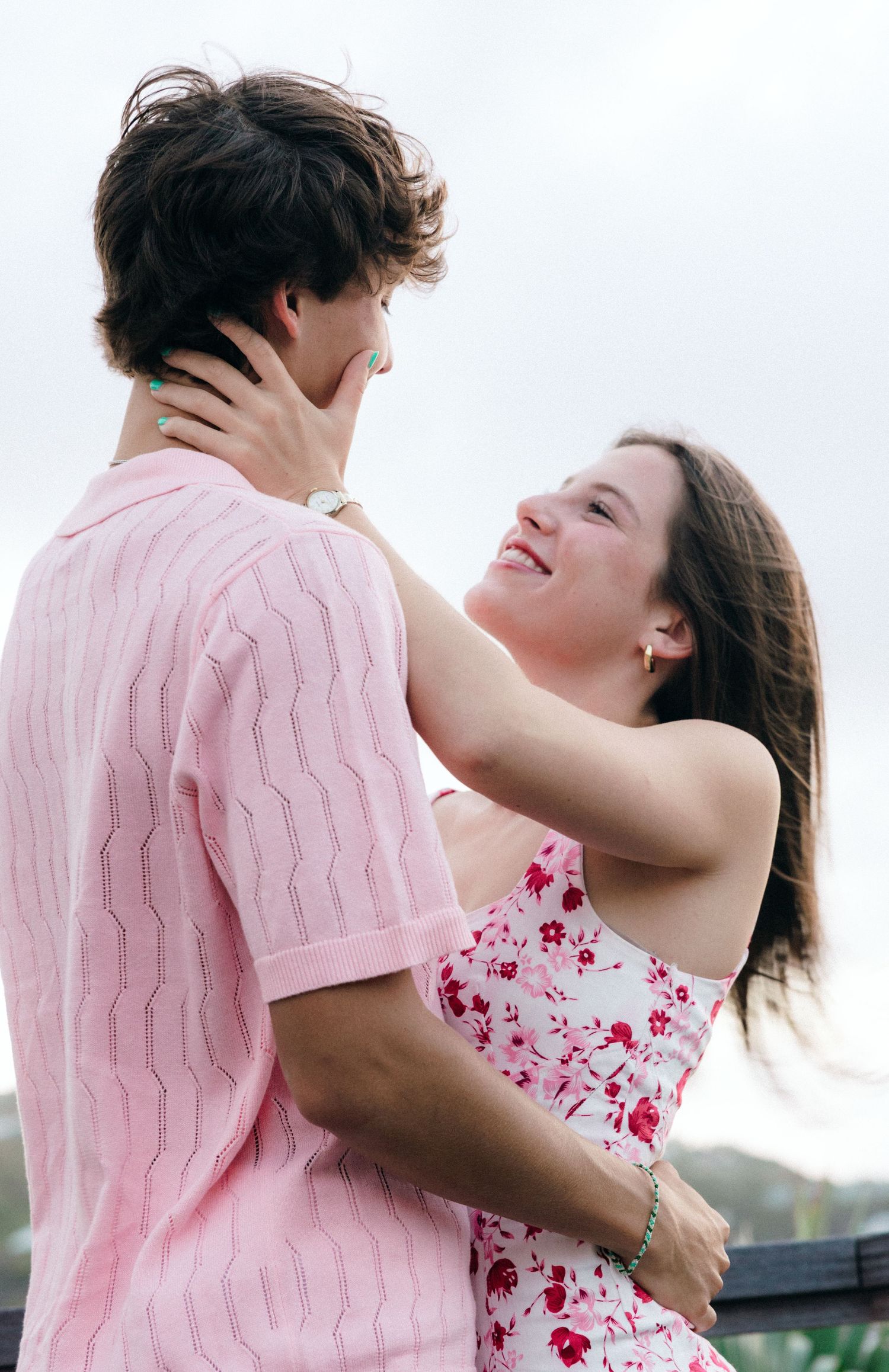 A smiling young couple in pink outfits share a romantic embrace outdoors, with the woman gazing up lovingly.