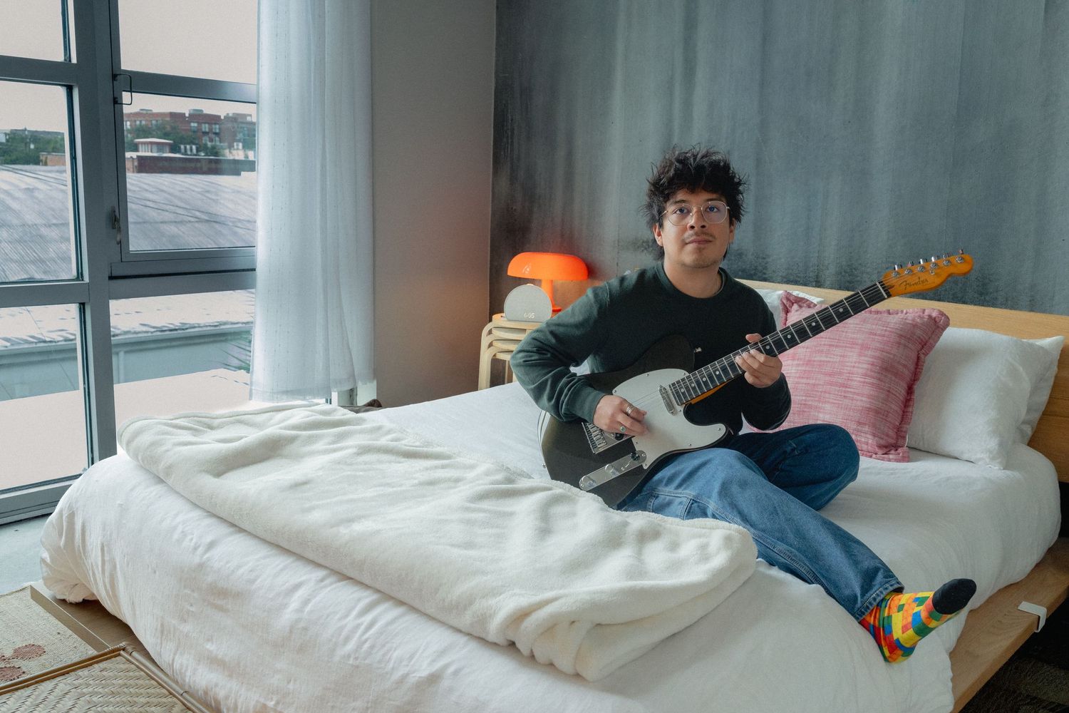 Young man with curly hair playing electric guitar while sitting on a white bed in a modern bedroom.