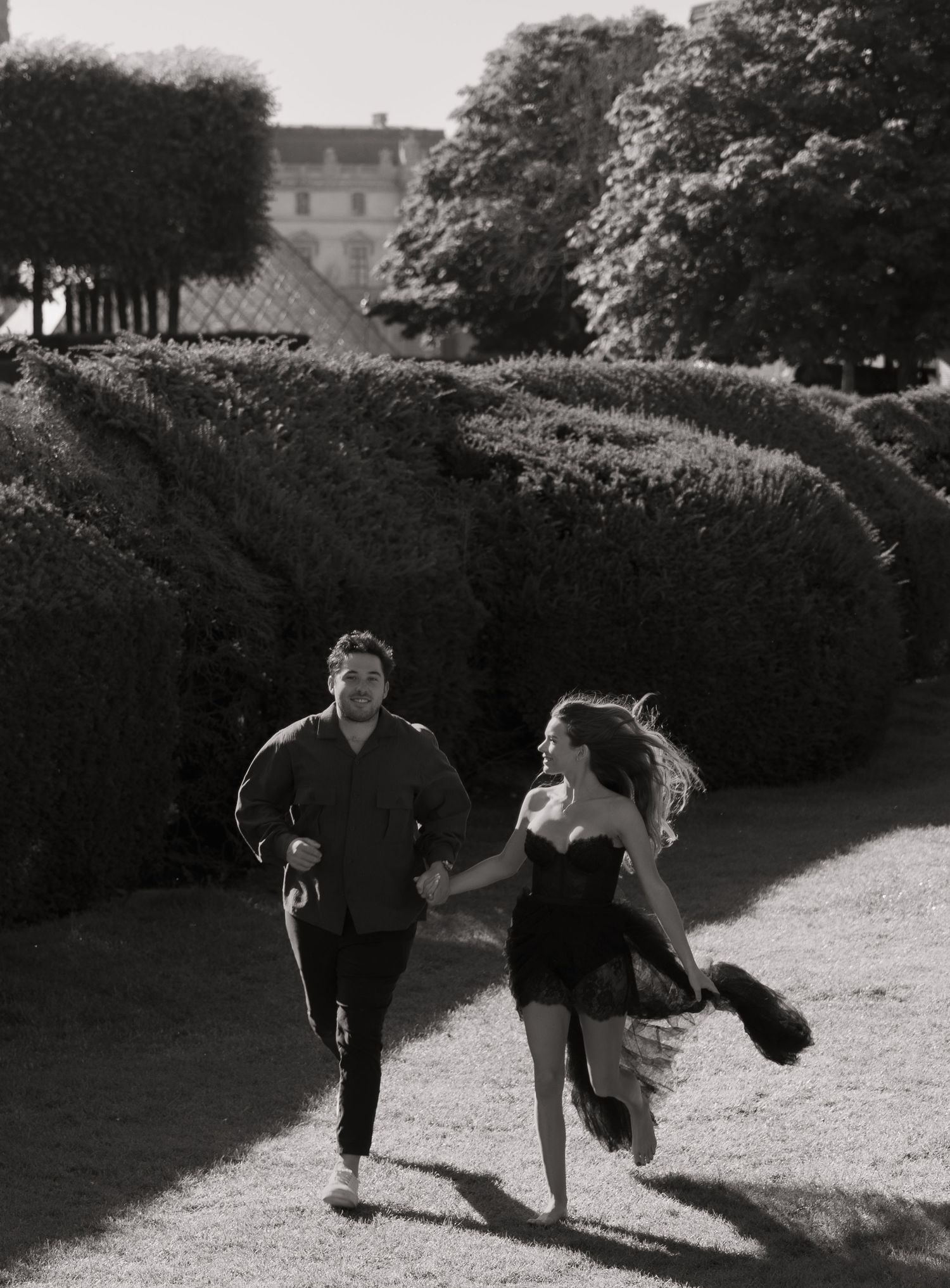 Black and white photo of joyful couple running hand in hand through a manicured garden.