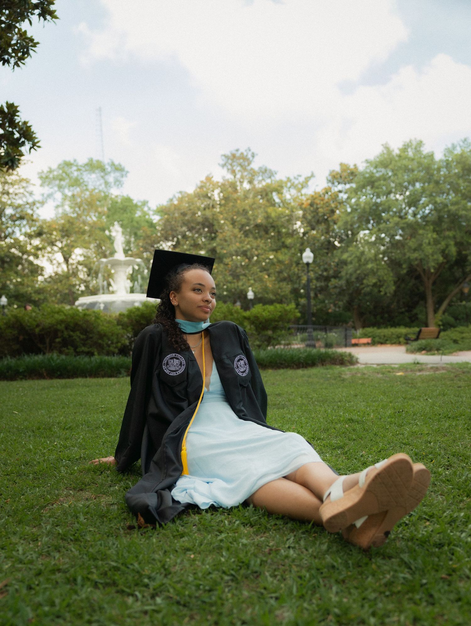 Female graduate in black cap and gown sits on grass near decorative fountain in park.