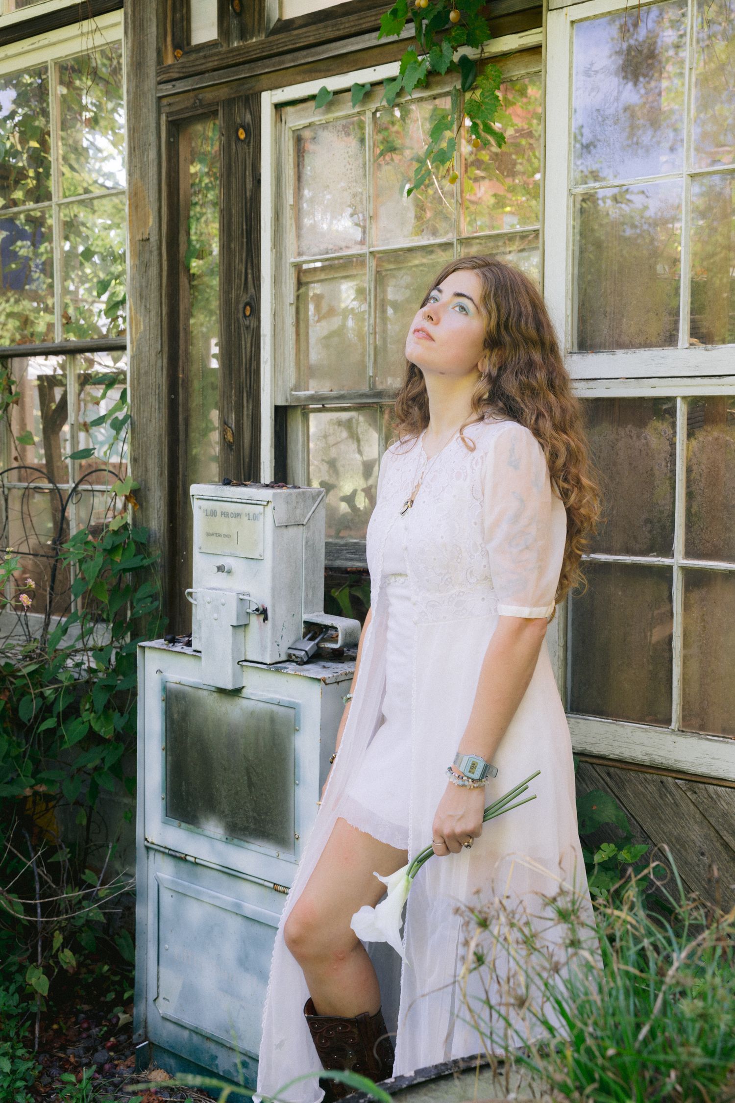 Young woman in white dress and brown boots holding flowers beside vintage appliance near rustic greenhouse windows.