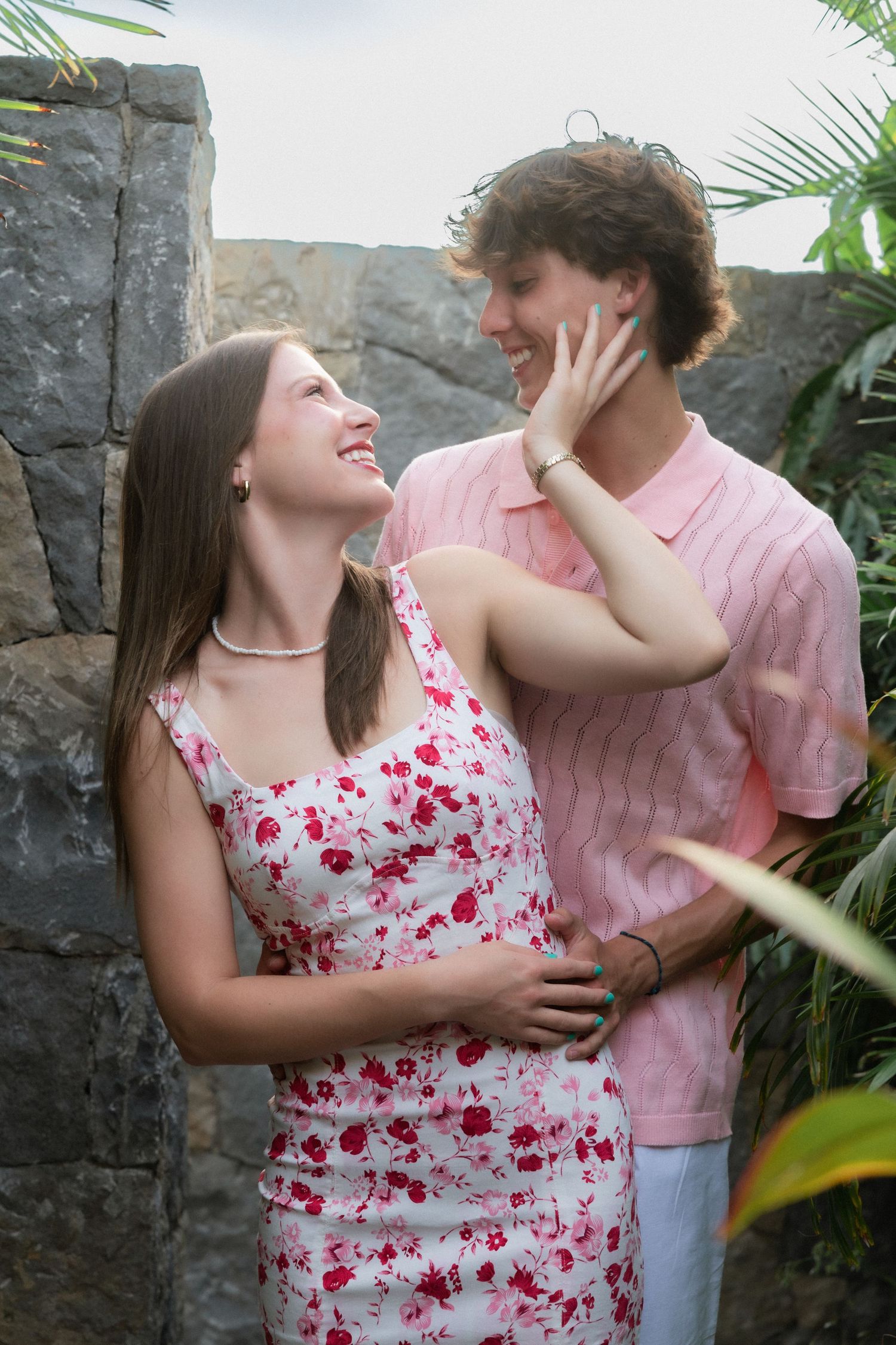 A young couple in pink and floral outfits share a romantic moment outdoors near stone walls and tropical plants.