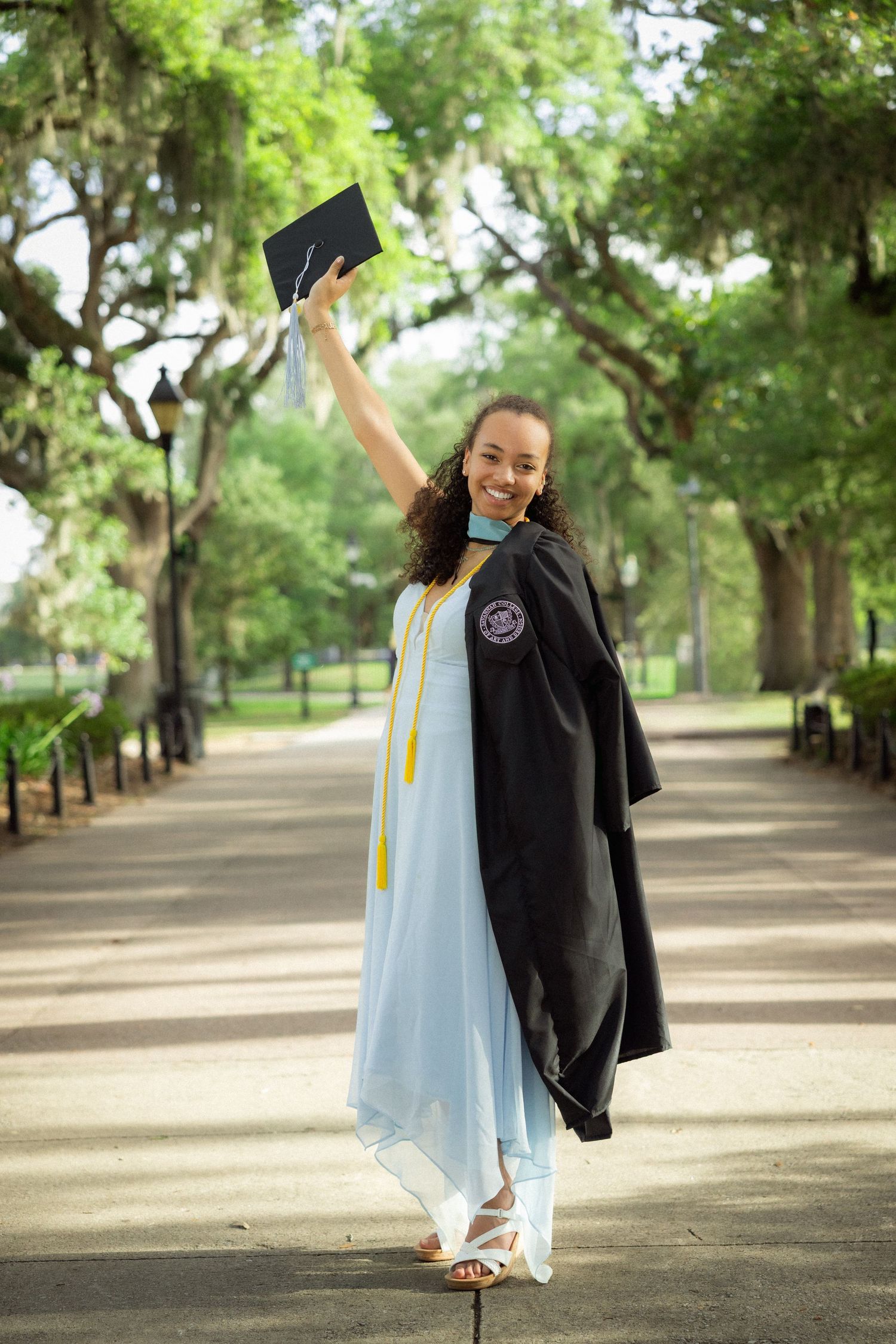 Smiling graduate in black gown raises cap in tree-lined park path.