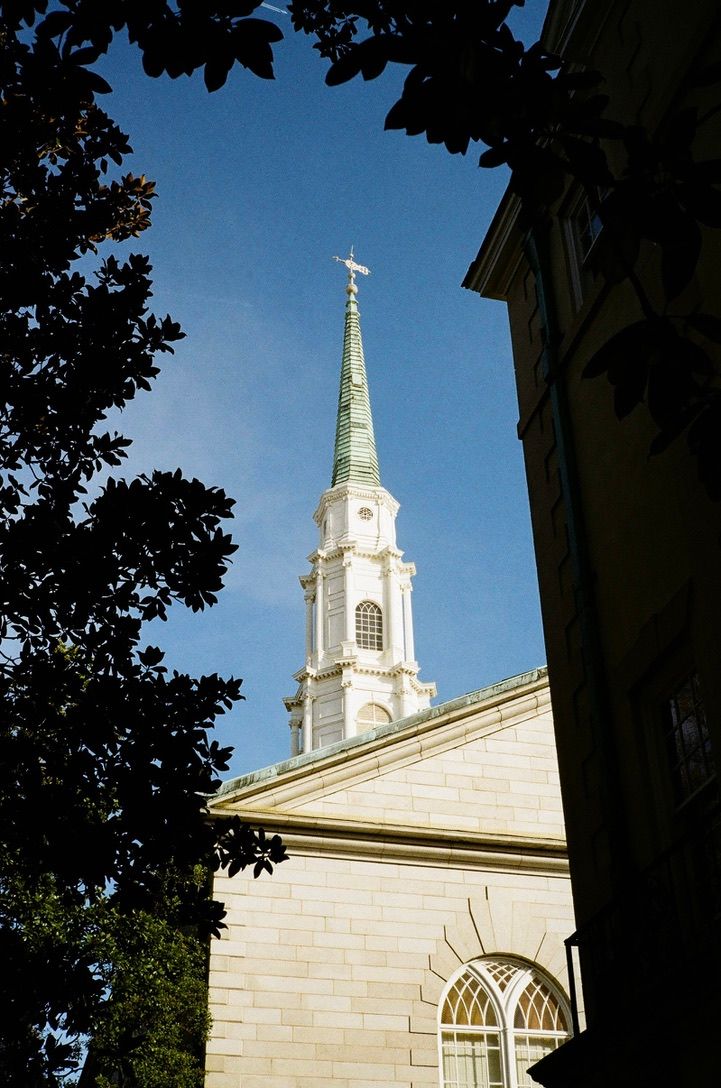 White church steeple with green spire and cross rises against blue sky, framed by trees and historic stone building.