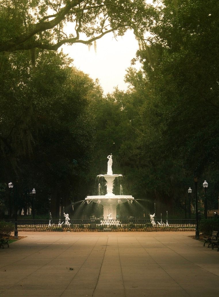 Ornate white tiered fountain surrounded by lush green trees in a serene park setting during golden hour.