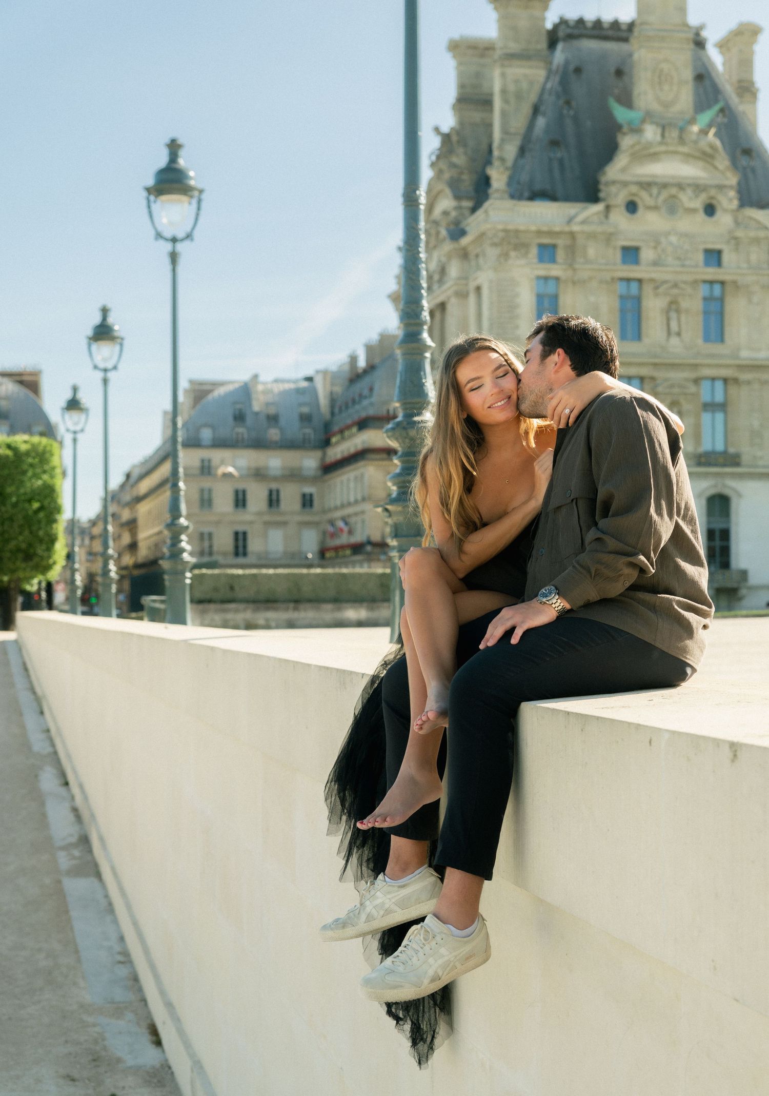 Couple sitting intimately on a Parisian wall near the Louvre with classic street lamps in background.