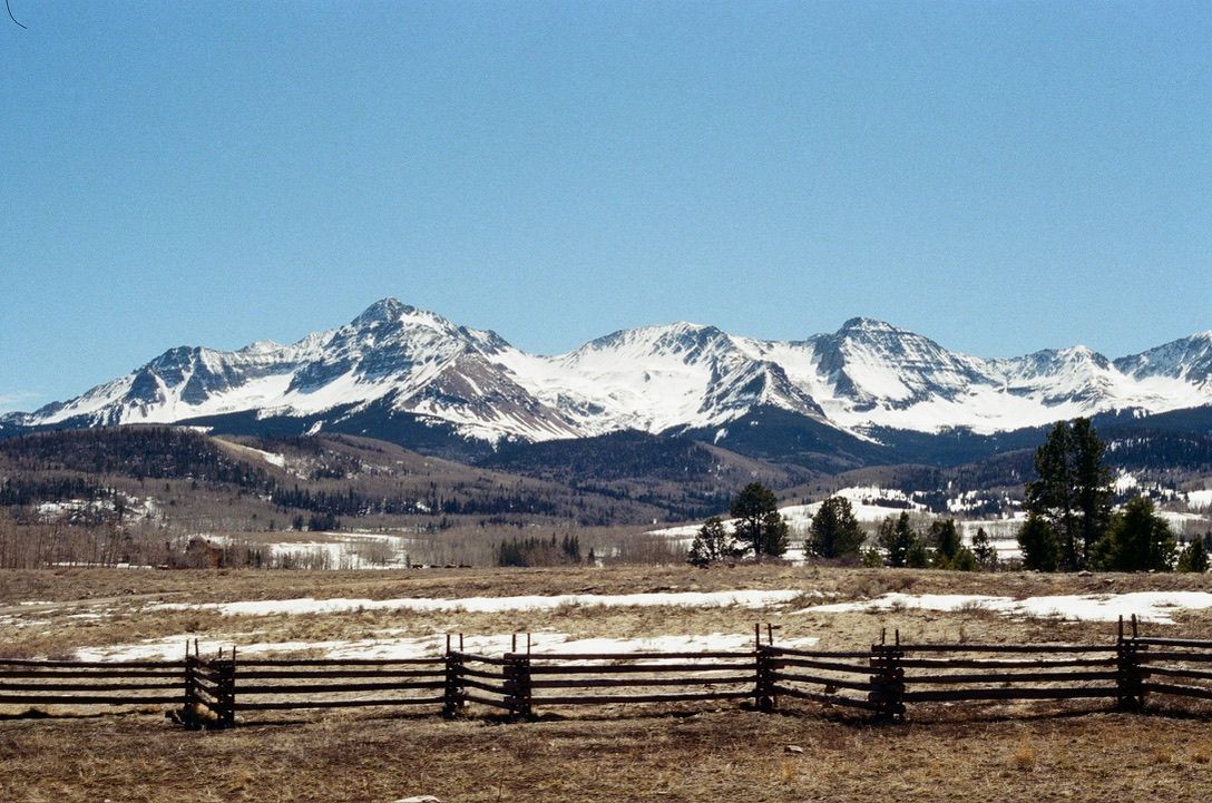 Snow-capped mountain range with wooden split-rail fence and open field in early spring.