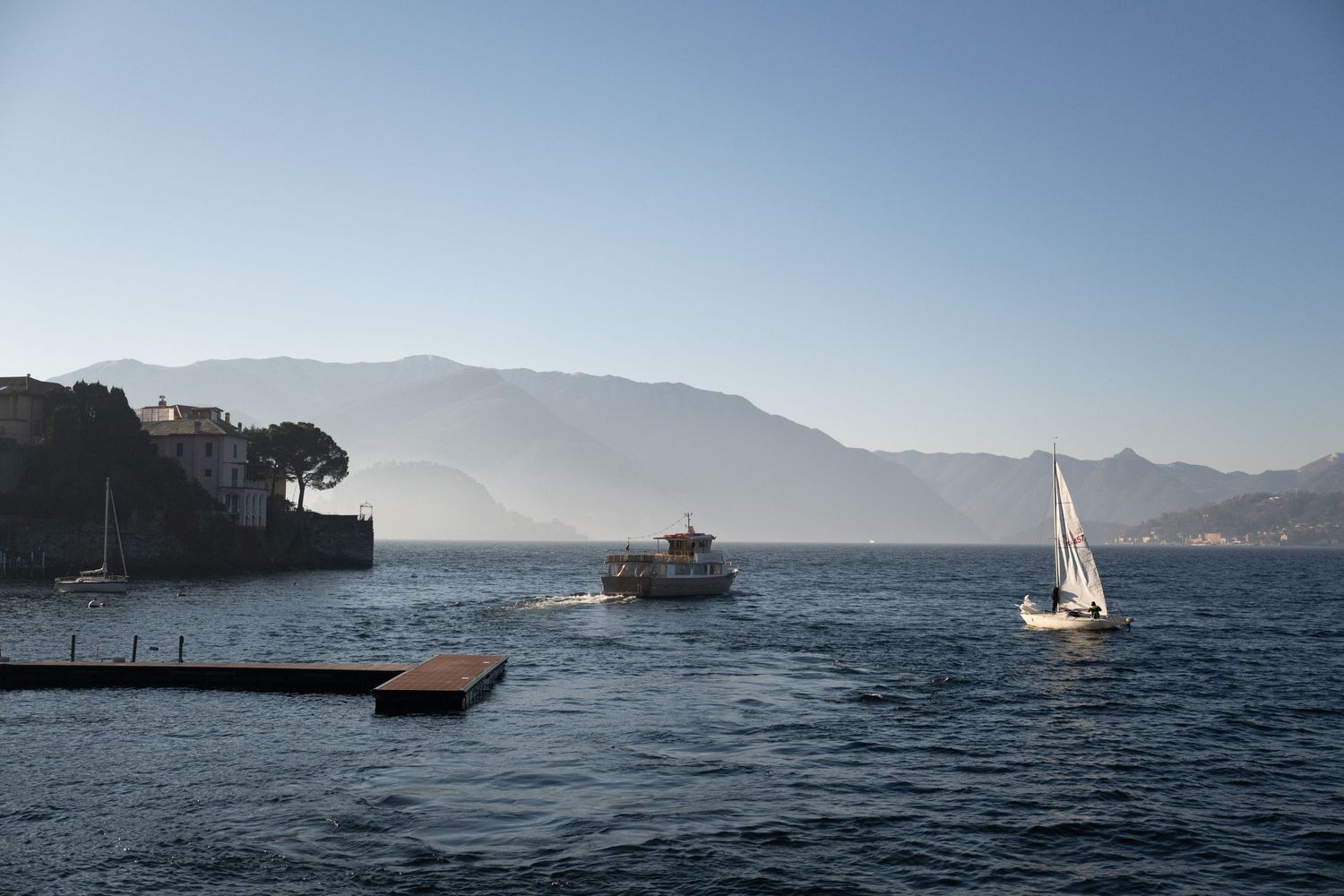 Sailboat and ferry on calm blue lake with misty mountains and historic village shoreline under clear sky.