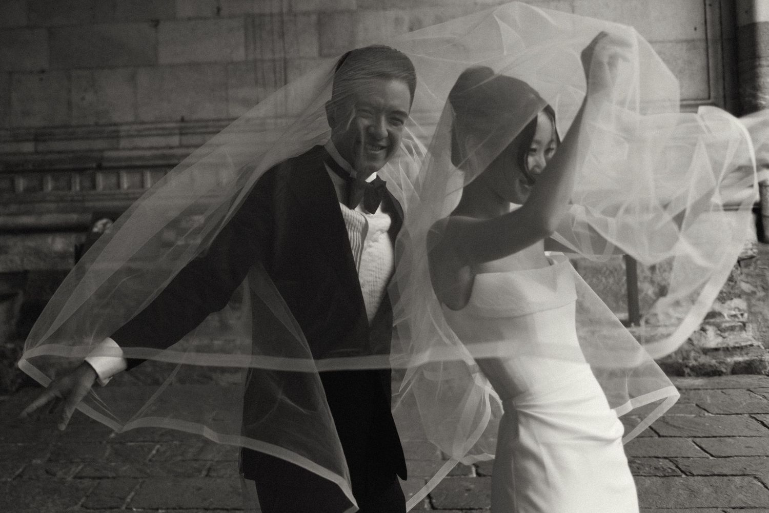 Black and white photo of a laughing bride and groom with a flowing veil billowing around them outdoors.