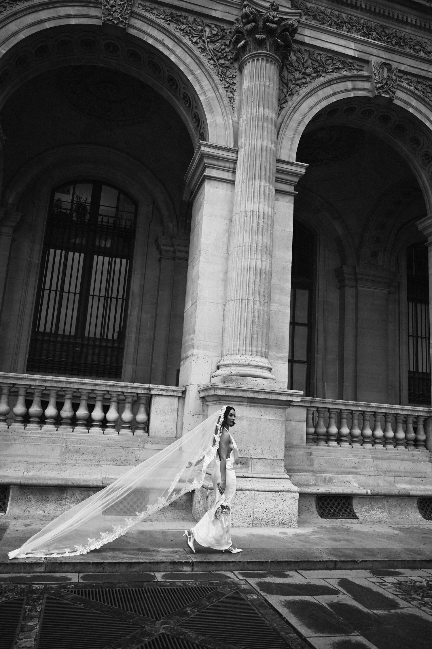 Elegant bride in sleek gown with dramatic lace veil flowing beside ornate arched columns of a classical building. Paris