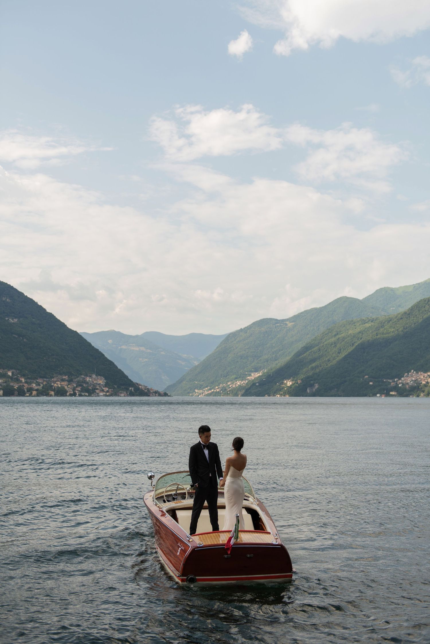 Bride and groom standing on a classic wooden Riva boat on scenic Lake Como surrounded by mountains.