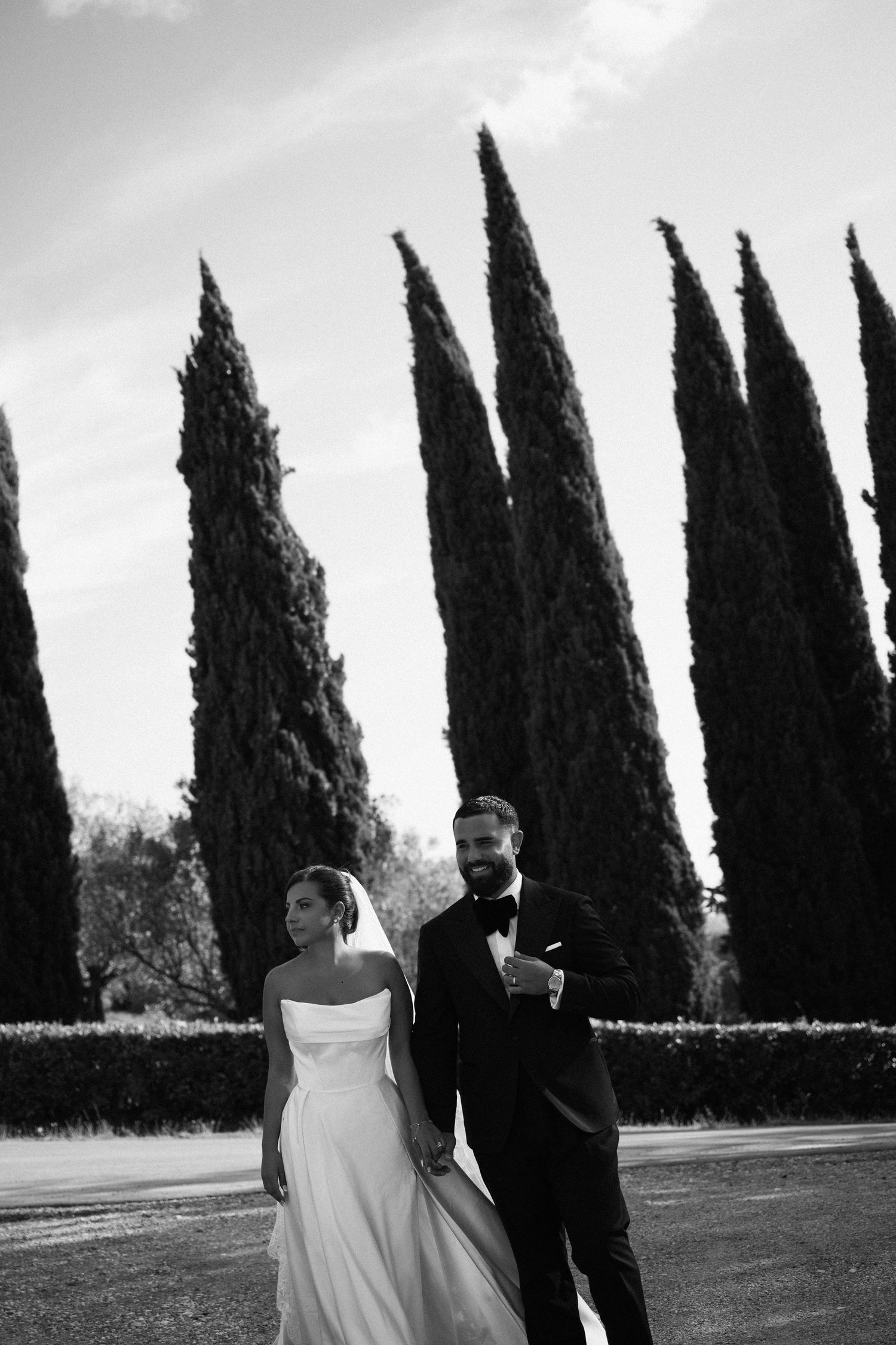 Bride and groom walk hand in hand before tall Italian cypress trees in elegant black and white portrait.