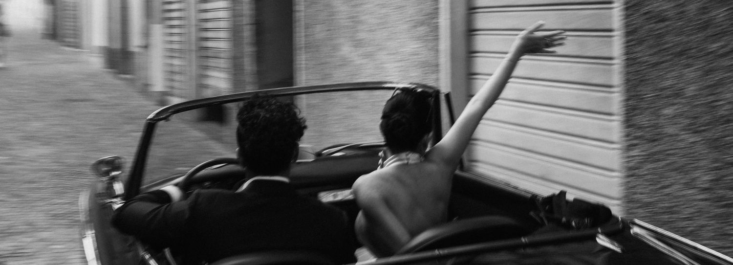 Black and white photo of a couple riding in a convertible car, woman raising her arm joyfully.