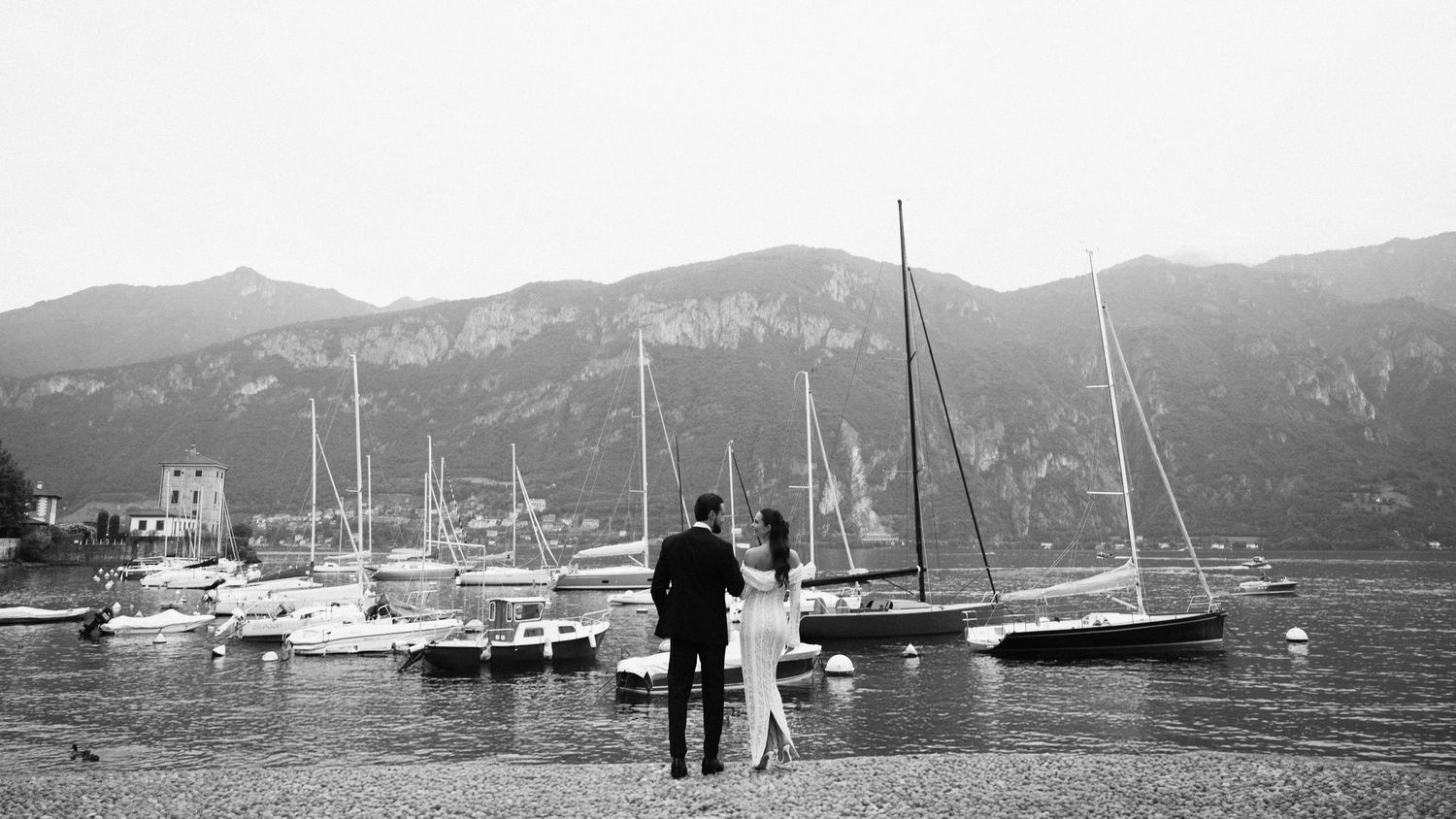 Bride and groom stand by a lakeside marina with sailboats and mountain backdrop in black and white wedding photo.