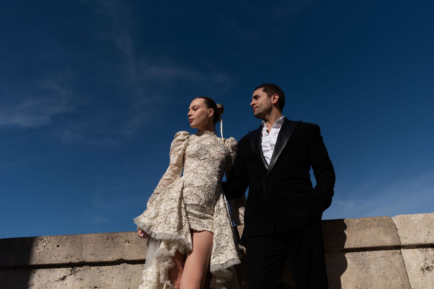 Stylish couple in lace mini dress and black suit posing against vivid blue Roman sky.