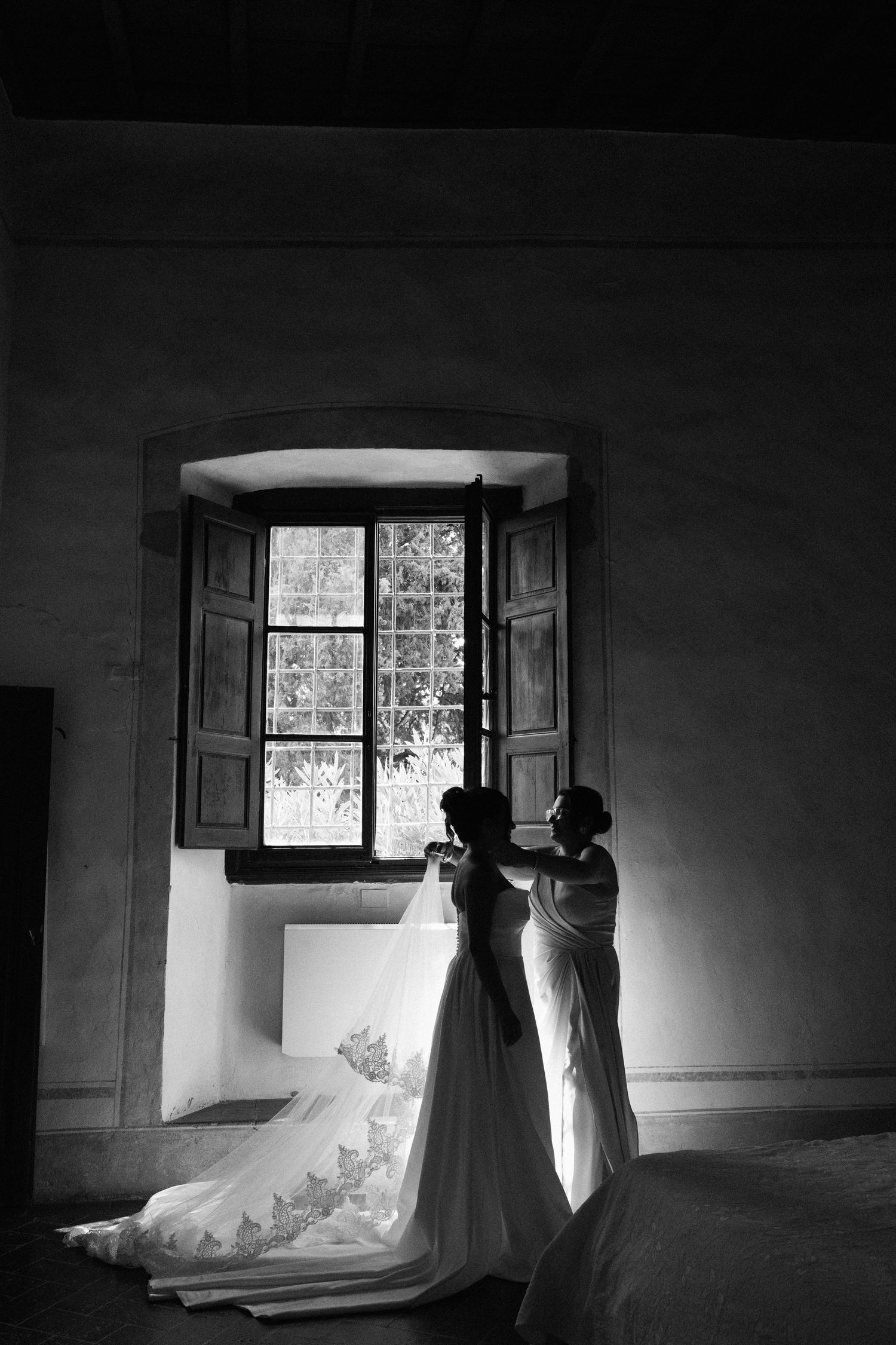 Bride being helped into wedding gown by silhouette of assistant near rustic arched window in black and white.