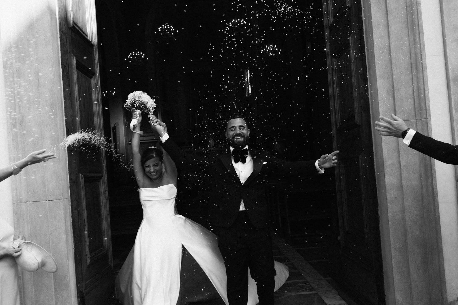 Newlyweds exit church doors in black and white as guests celebrate with confetti shower.