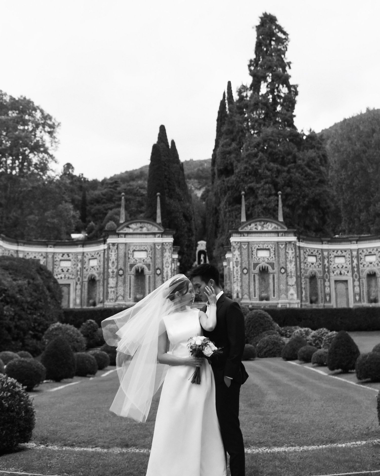 Black and white photo of bride and groom kissing in ornate Italian garden with cypress trees. Villa d'Este Lake Como