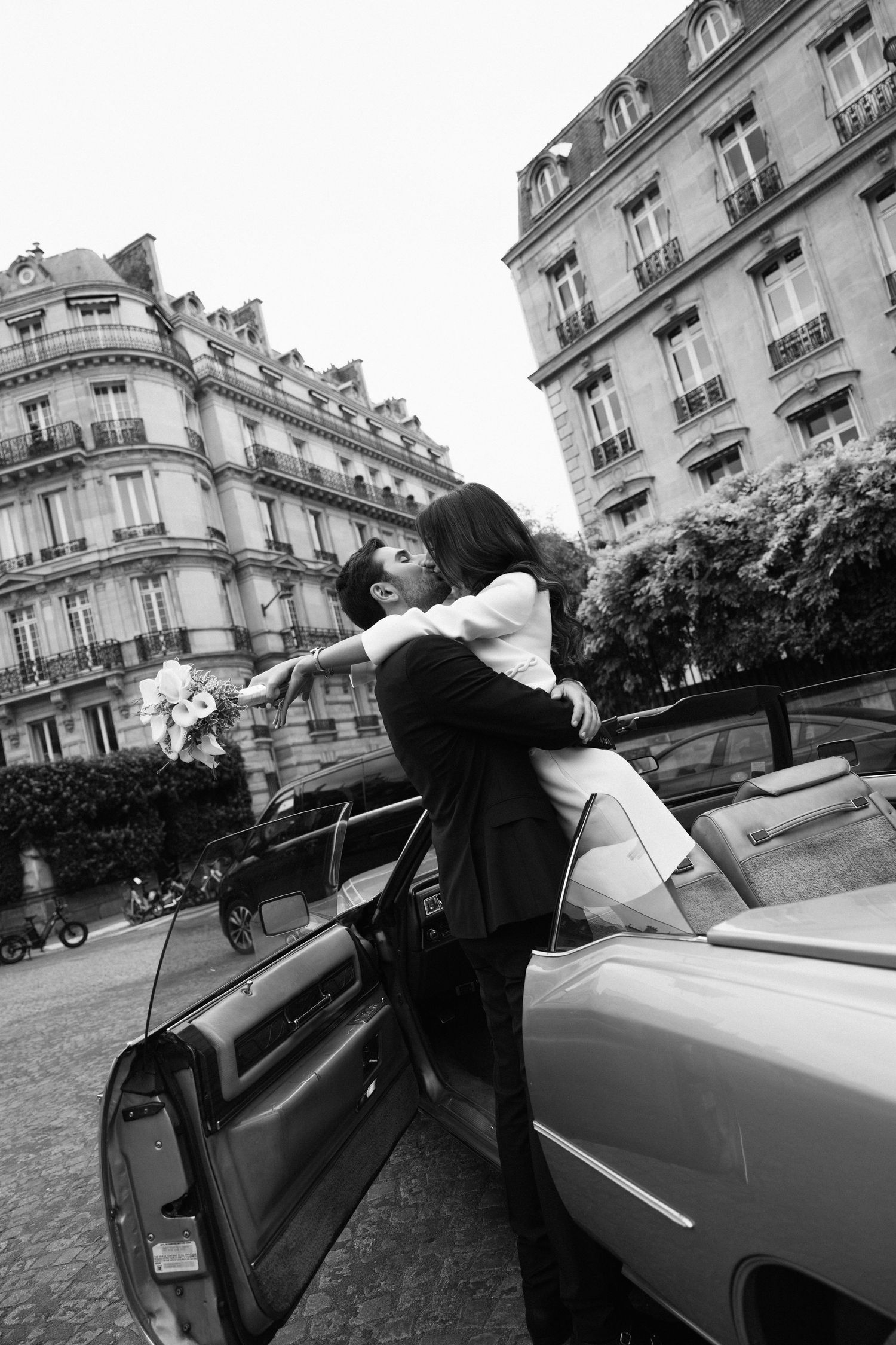 Black and white photo of a groom lifting his bride beside a vintage convertible on a Paris street.