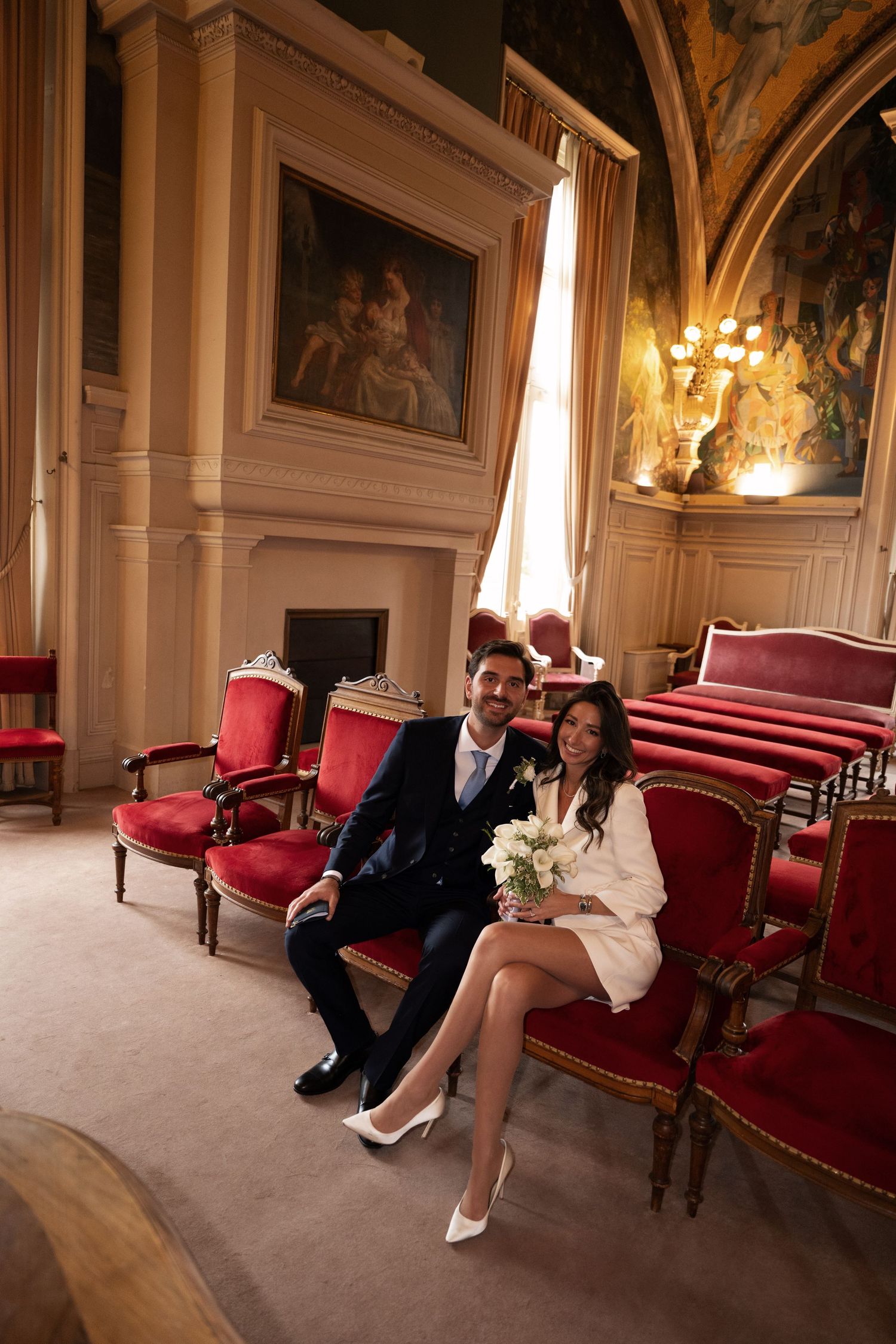 Elegant wedding couple sitting on red velvet chairs in a grand ornate French château interior.