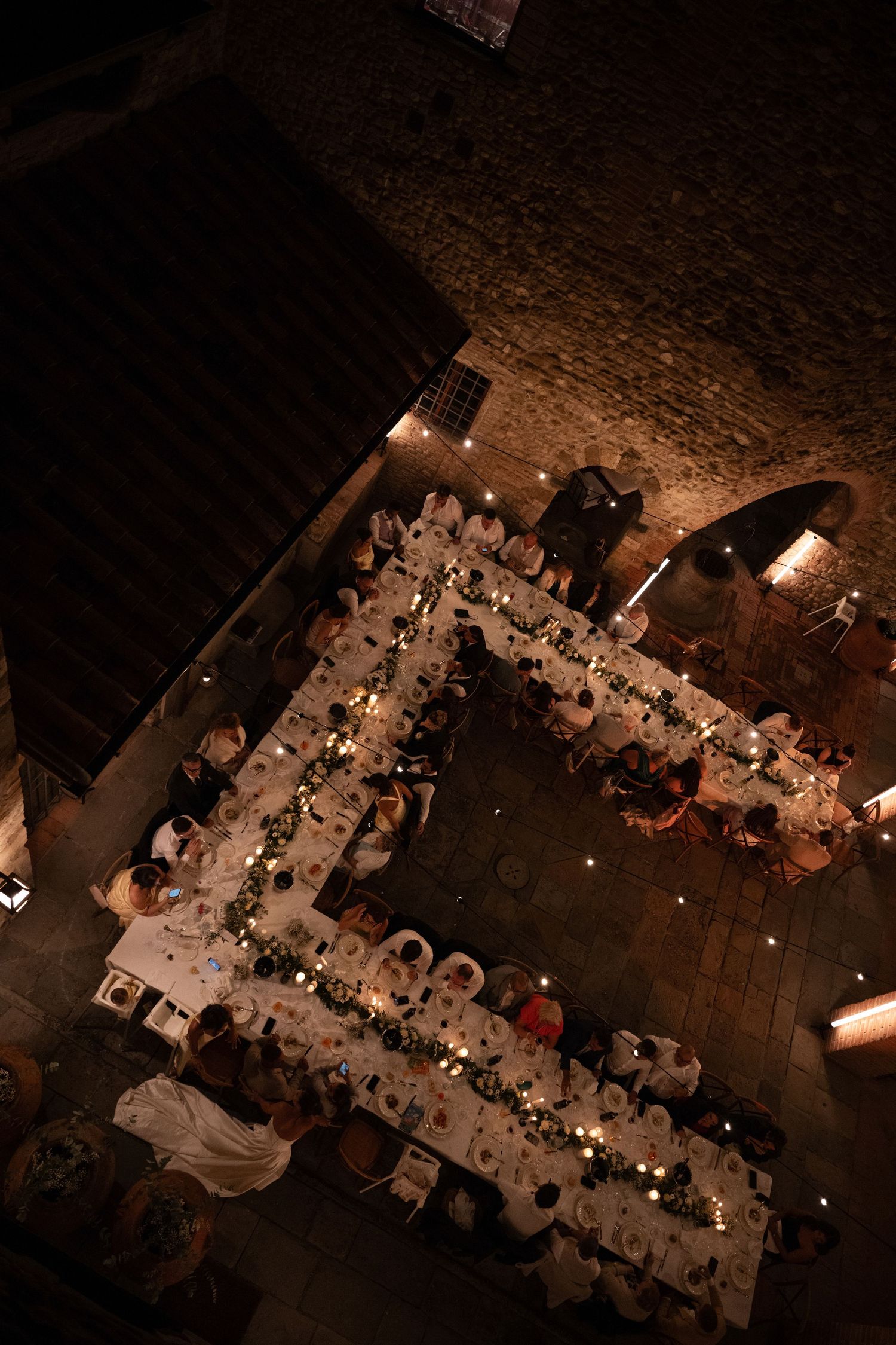 Bird's eye view of intimate candlelit wedding reception with greenery-adorned tables in a historic stone courtyard.