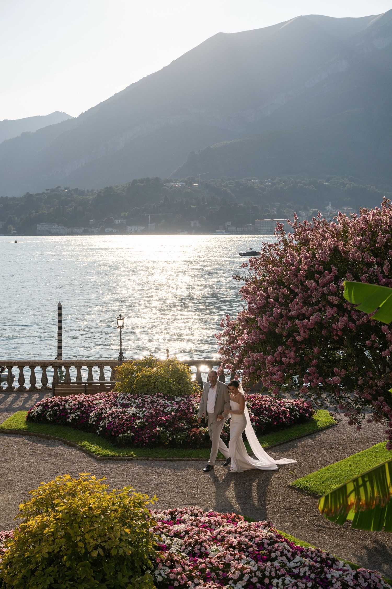 Bride and groom walking through colorful garden with lake and mountains backdrop at golden hour. Lake Como