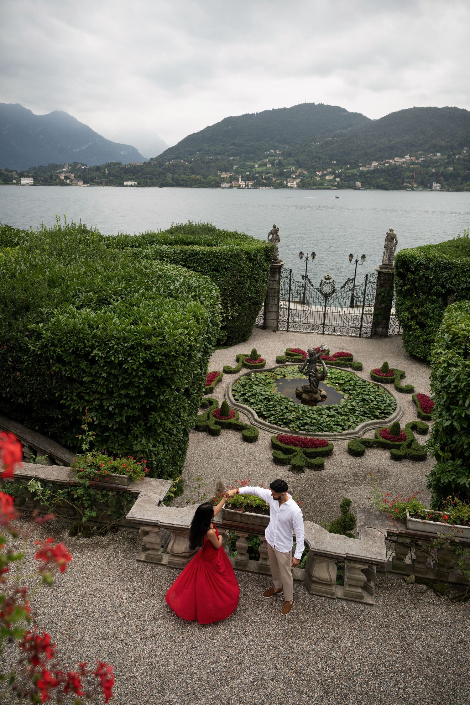 Aerial view of couple in red dress exploring ornate Italian villa garden with lake backdrop. Lake Como