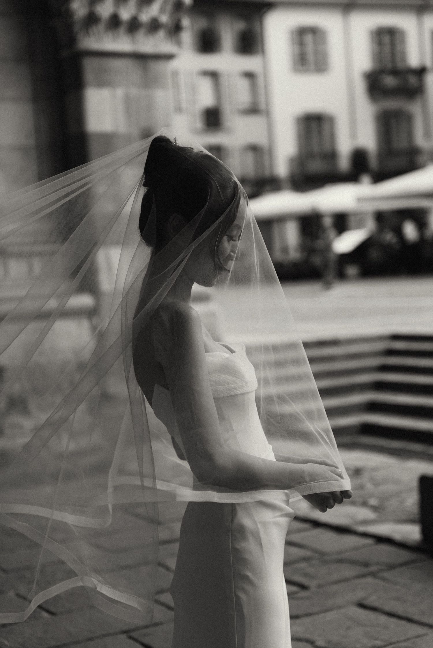 Black and white portrait of a bride in a sleek wedding dress with a flowing veil in an Italian piazza.