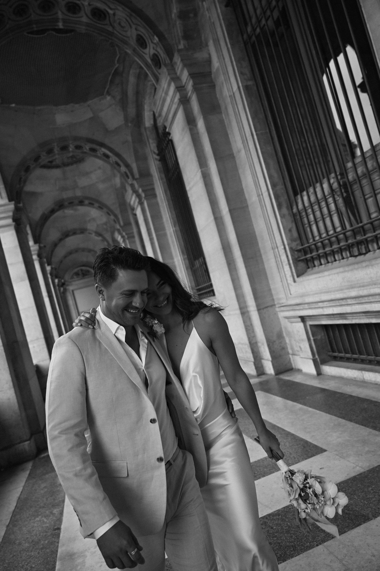Black and white photo of laughing bride and groom walking together through sunlit classical architectural colonnade. Paris