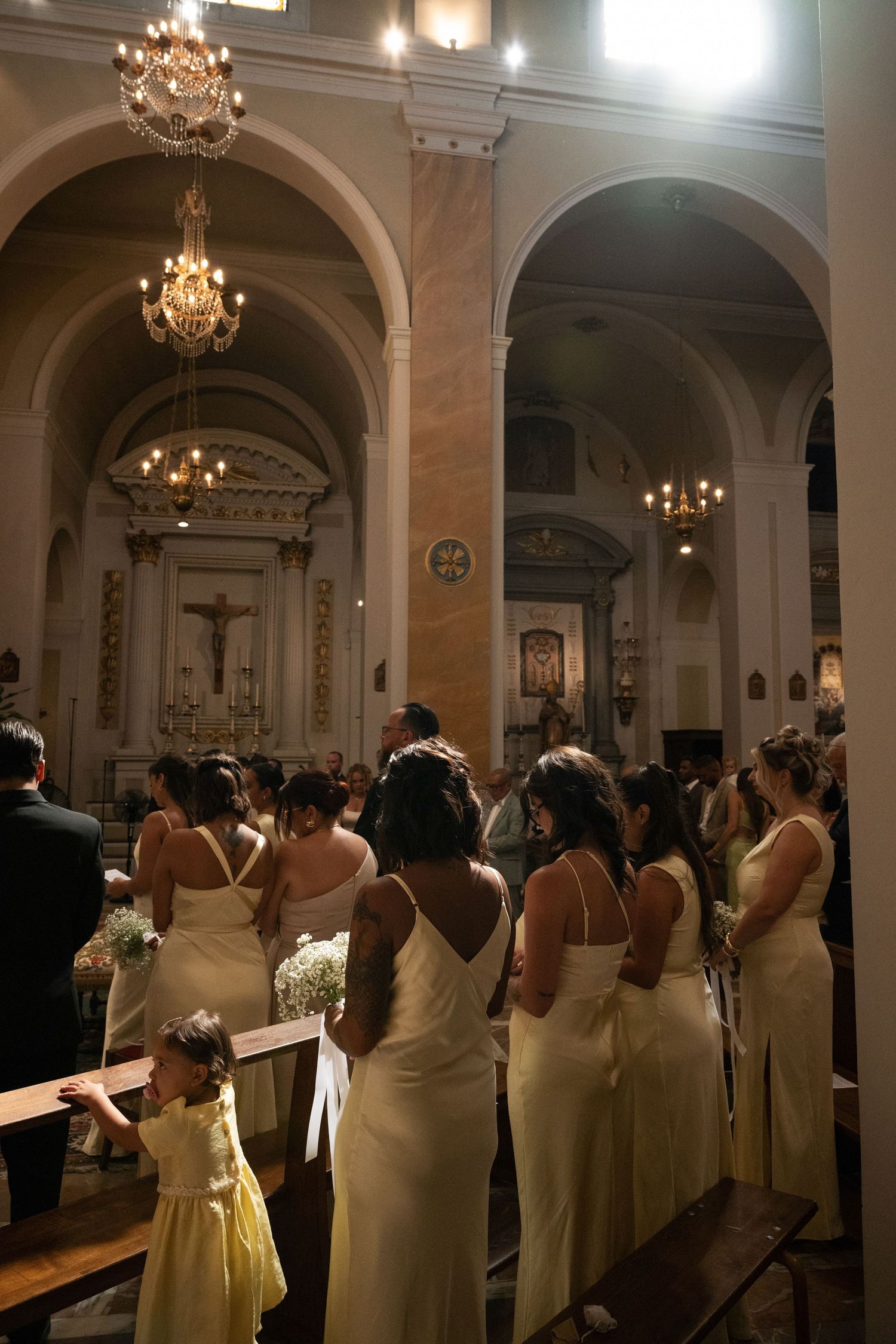 Bridesmaids in ivory gowns stand in ornate church aisle during wedding ceremony with chandeliers above.
